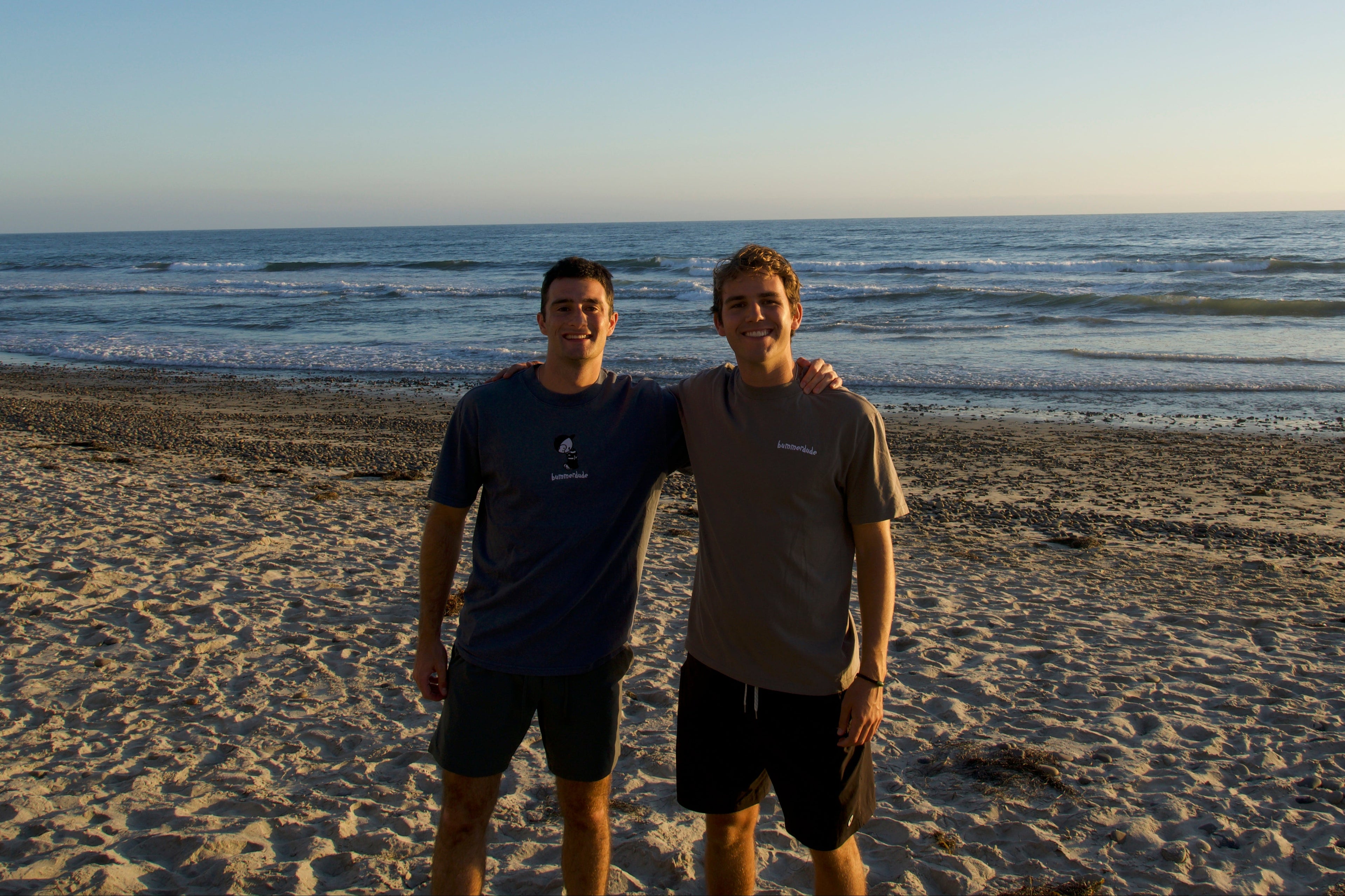 Two people standing on a beach with ocean in the background wearing bummerdude